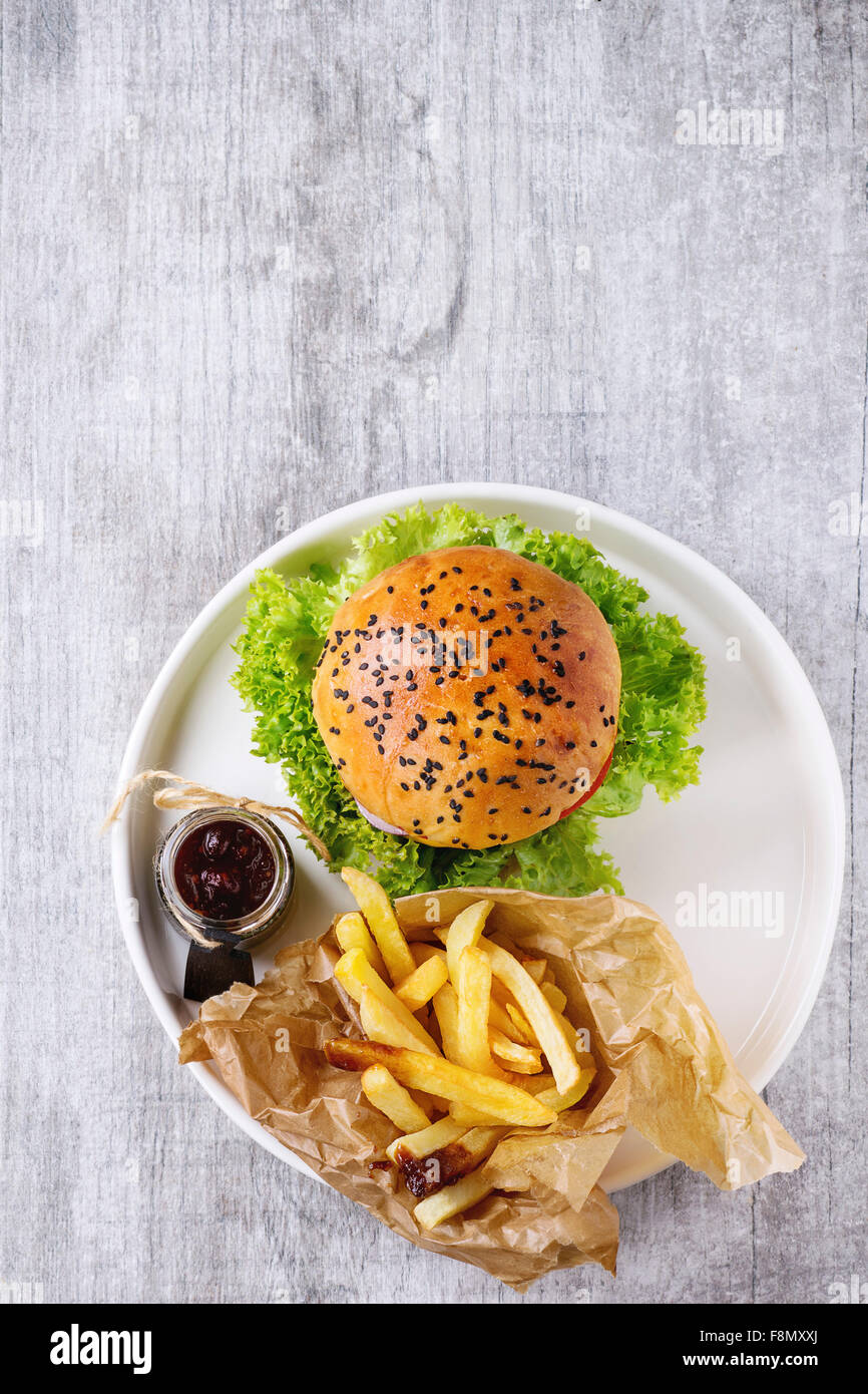 Frische hausgemachte Burger mit schwarzem Sesam in weißen Teller mit Bratkartoffeln, serviert mit Ketchup-Sauce im Glas über gr Stockfoto