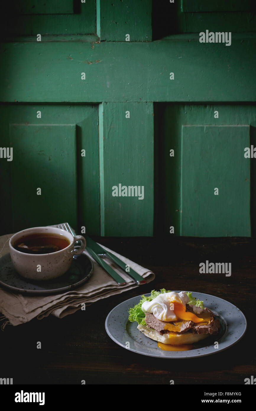 Café-Frühstück mit hausgemachten Sandwich mit gebackenem Fleisch und weich gekochtes Ei und heiße Tasche Tee über dunklen Holztisch in der Nähe Stockfoto