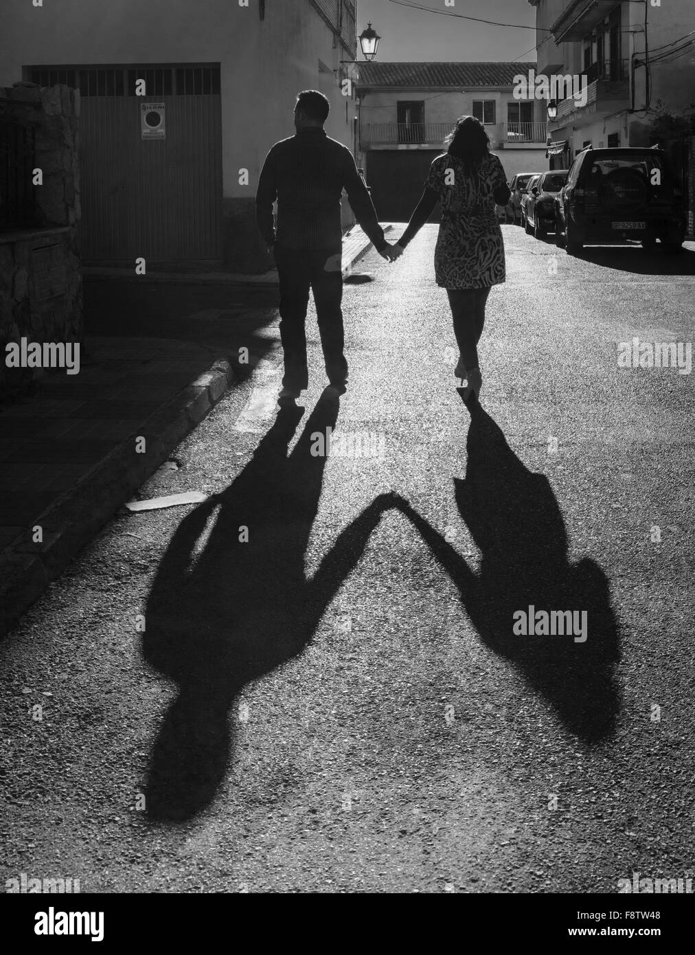 Junge Liebhaber gehen Hand in Hand mit dem Rücken auf der Straße, die Kamera und die langen Schatten der Silhouette dahinter. Stockfoto