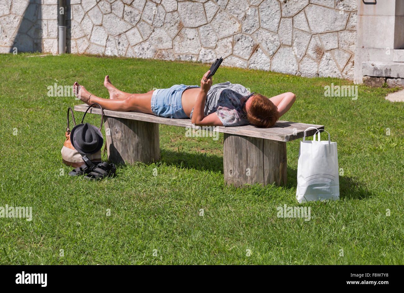 Kaukasische Frau mittleren Alters in Shorts und Sonnenbrille liegen nach dem Einkauf auf einer Bank im Freien und elektronische Lesebuch gegerbt Stockfoto