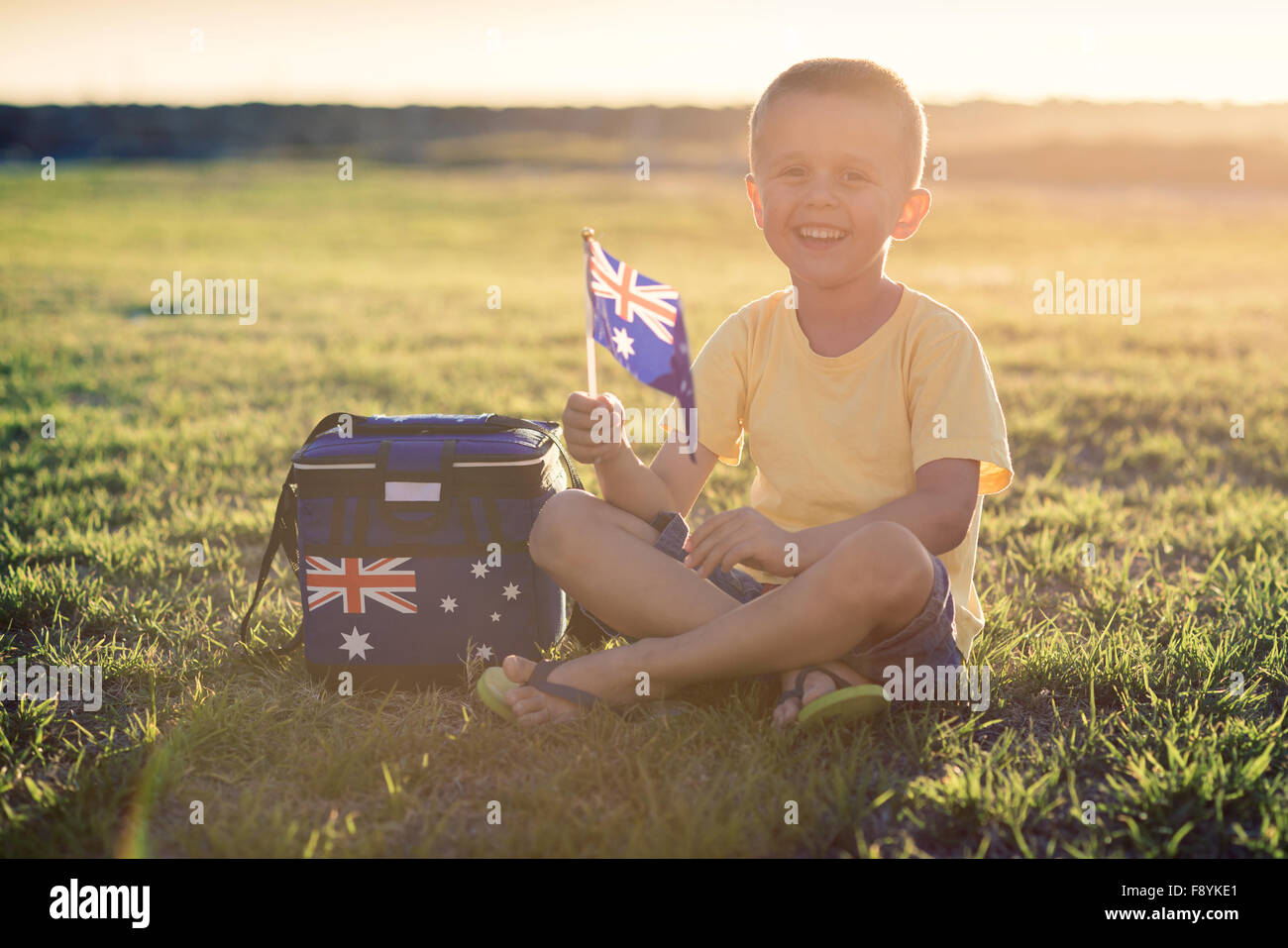 Custe lächelndes Kind mit Flagge Australiens sitzen auf dem Rasen bei Sonnenuntergang am Australia Day Stockfoto