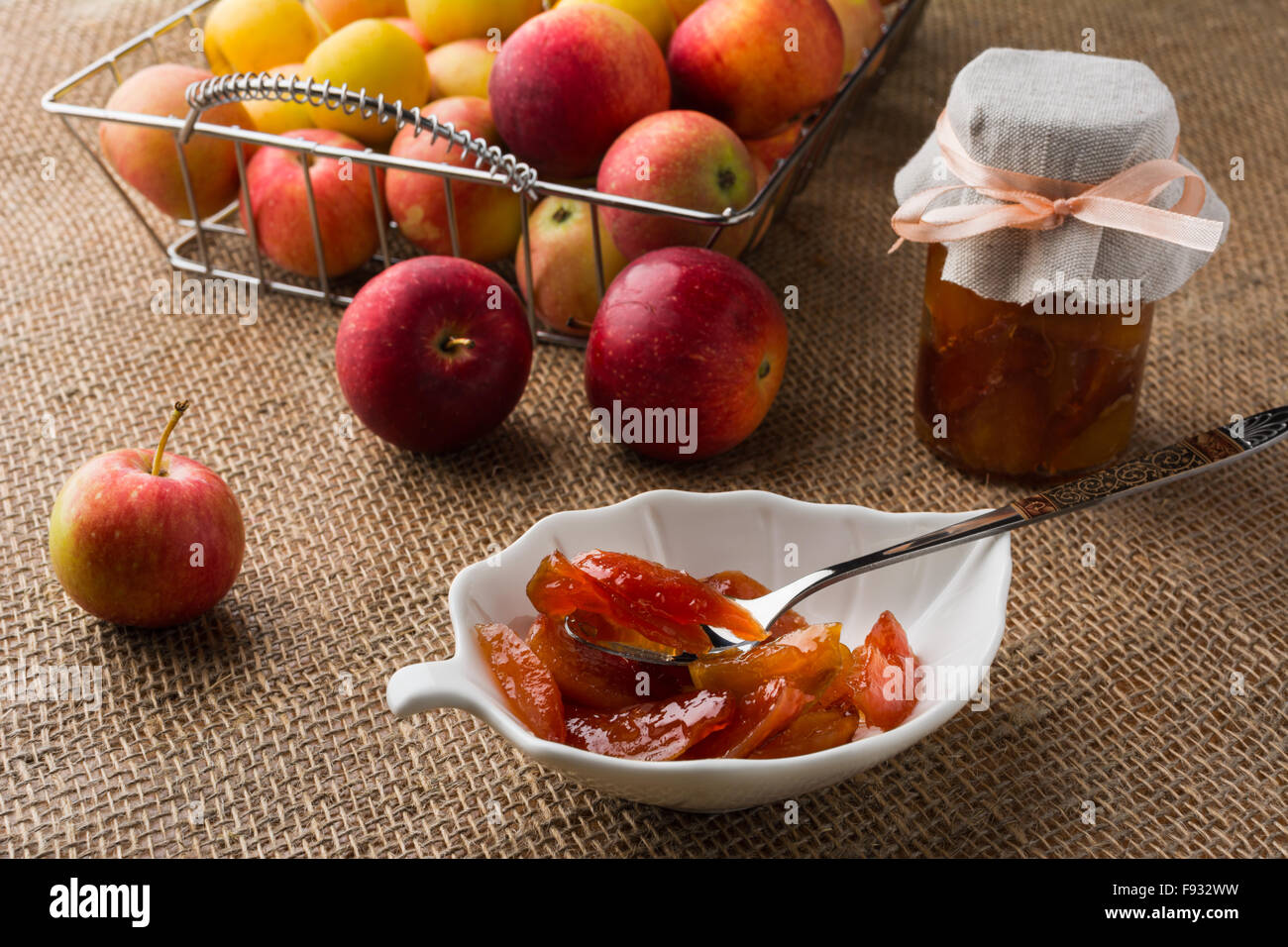 Hausgemachte Apfel Marmelade auf weiße Untertasse, Löffel, Glas Marmelade und Äpfel in Metall Drahtkorb auf einem Tisch mit Sackleinen abgedeckt. Sele Stockfoto