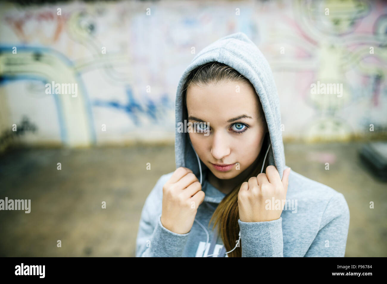 Schwere Teenager-Mädchen mit Kapuze auf anhören von Musik über Kopfhörer auf Hintergrund der Graffitiwand stehend Stockfoto