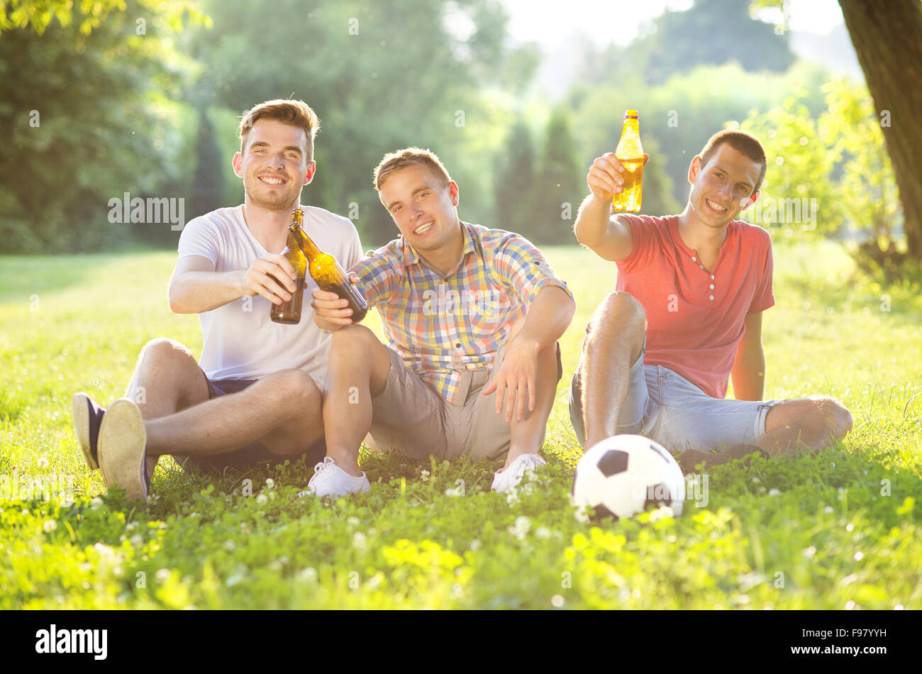 Drei glückliche Freunde verbringen Freizeit zusammen im Park sitzen auf dem Rasen, Bier trinken und plaudern Stockfoto