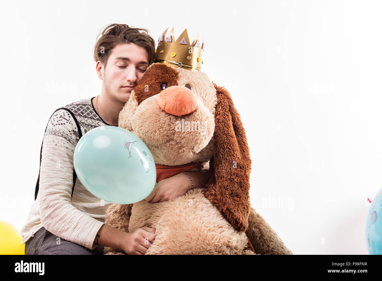 Junger Mann umarmt eine großer Hund Puppe schlafen auf einer Party, vielleicht mit einem Kater, der Hund hat eine Krone und es gibt Luftballons um Stockfoto