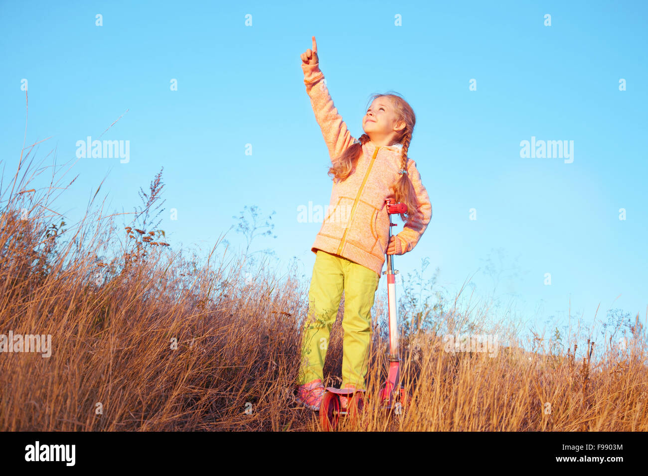 Kleine Mädchen in helle Kleidung mit Scooter zeigt einen Finger in den Himmel Stockfoto
