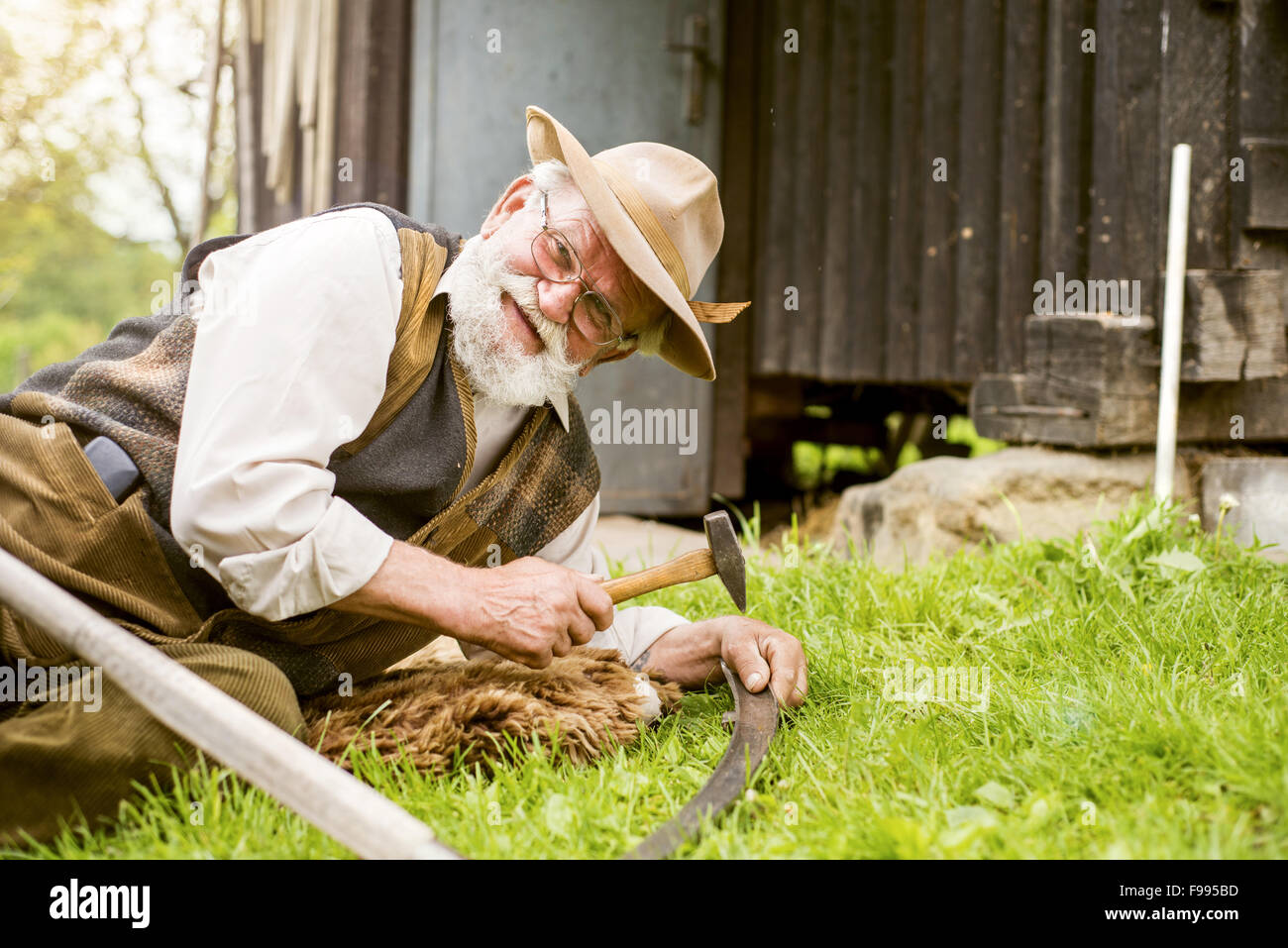 Alter Bauer mit Bart ist seine Sense reparieren, bevor Sie mit traditionell den Rasen mähen Stockfoto