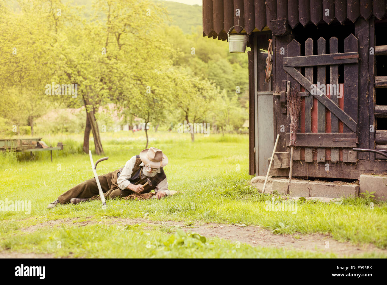 Alter Bauer mit Bart ist seine Sense reparieren, bevor Sie mit traditionell den Rasen mähen Stockfoto