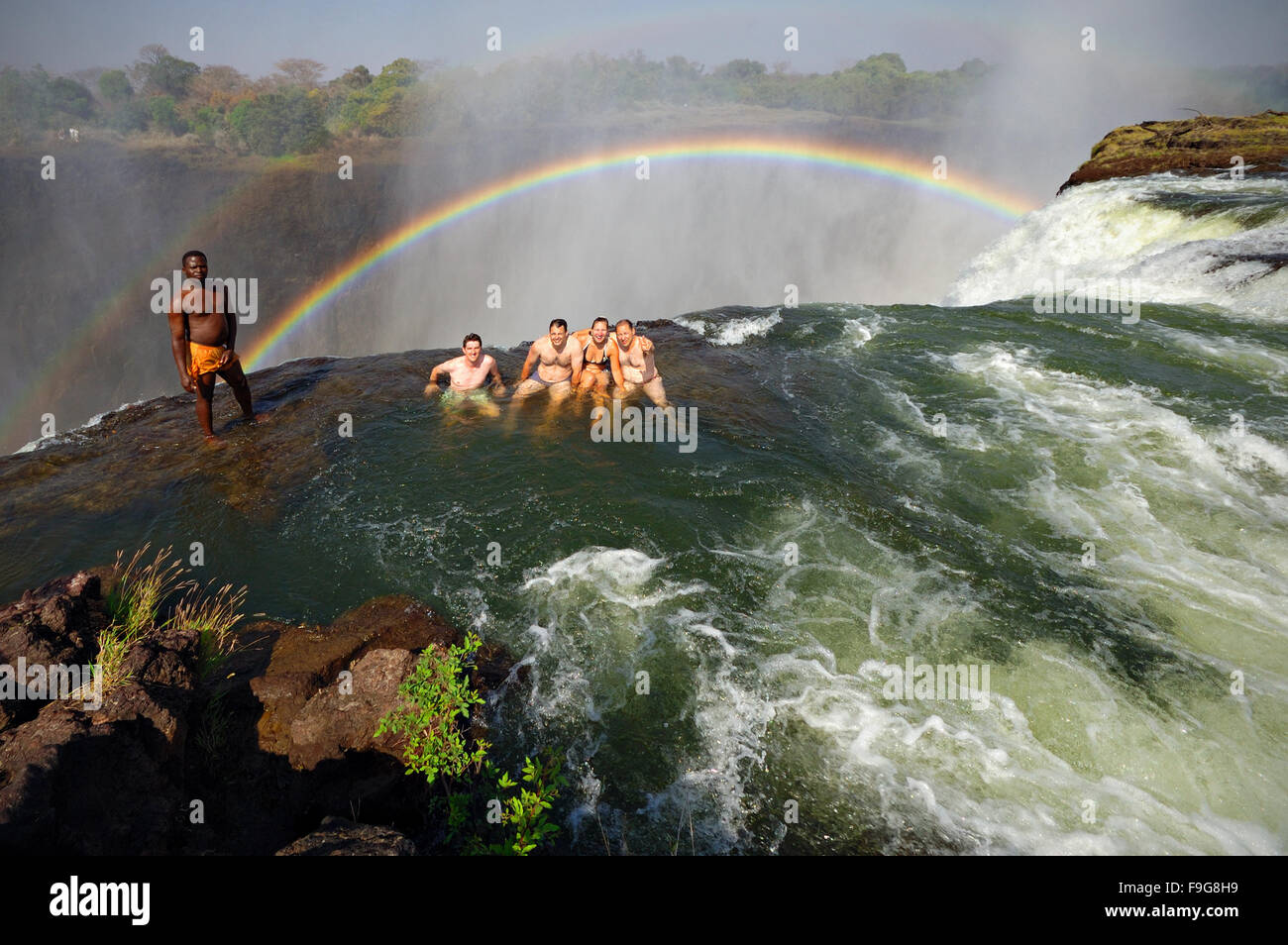 Vier westliche Touristen schwimmen am Rande der Victoria Falls in Devil's Pool unter der Aufsicht eines afrikanischen Führer, Sambia Stockfoto