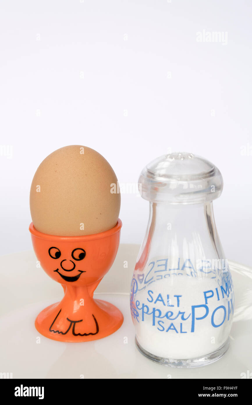 Eine orange Eierbecher mit einem gemalten Smiley-Gesicht, hält ein unversehrtes braune Ei neben einen Topf mit Salz Stockfoto