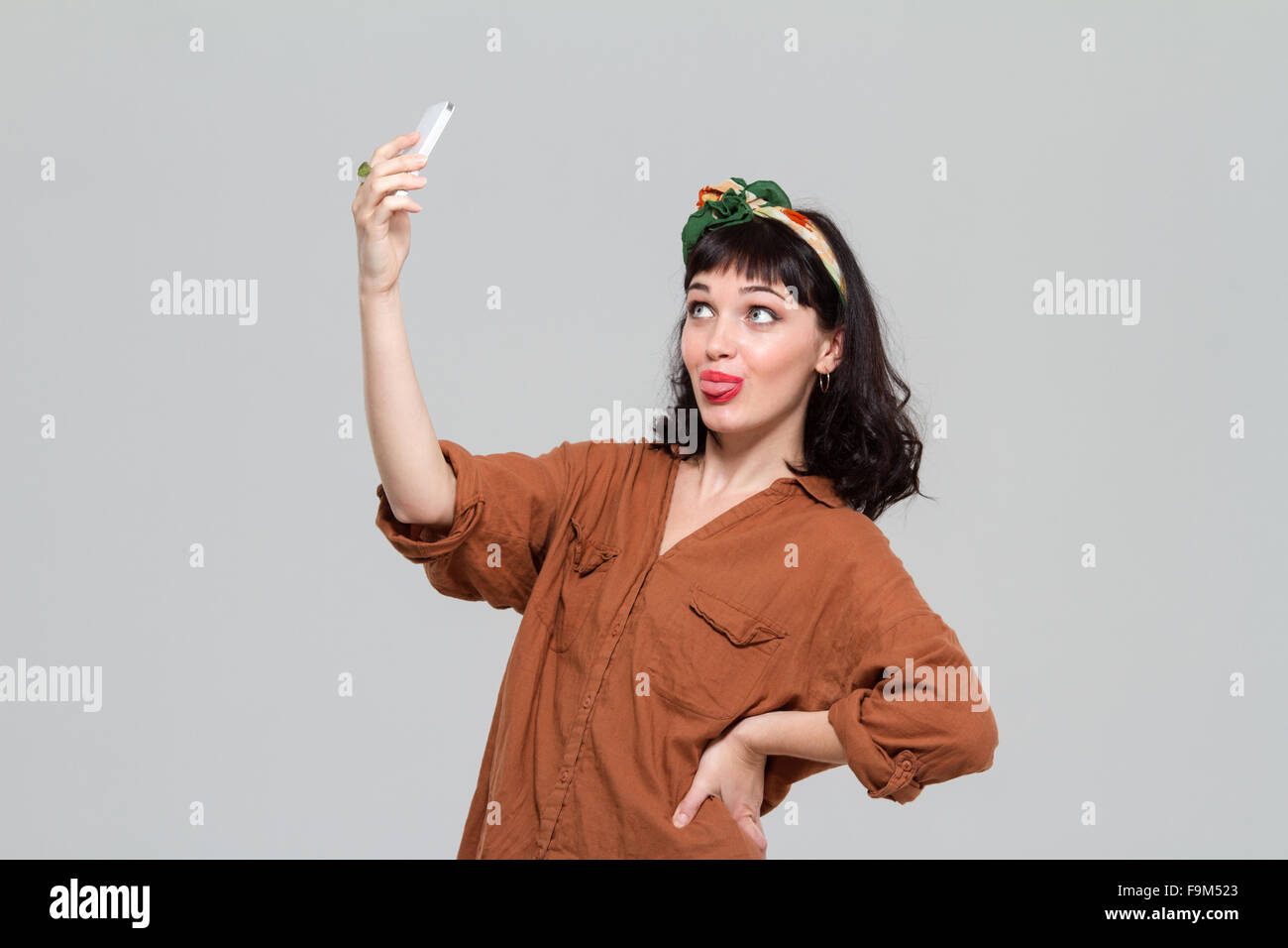 Amüsante fröhliche junge Frau unter Selfie und zeigt Zunge mit Smartphone über grauen Hintergrund isoliert Stockfoto
