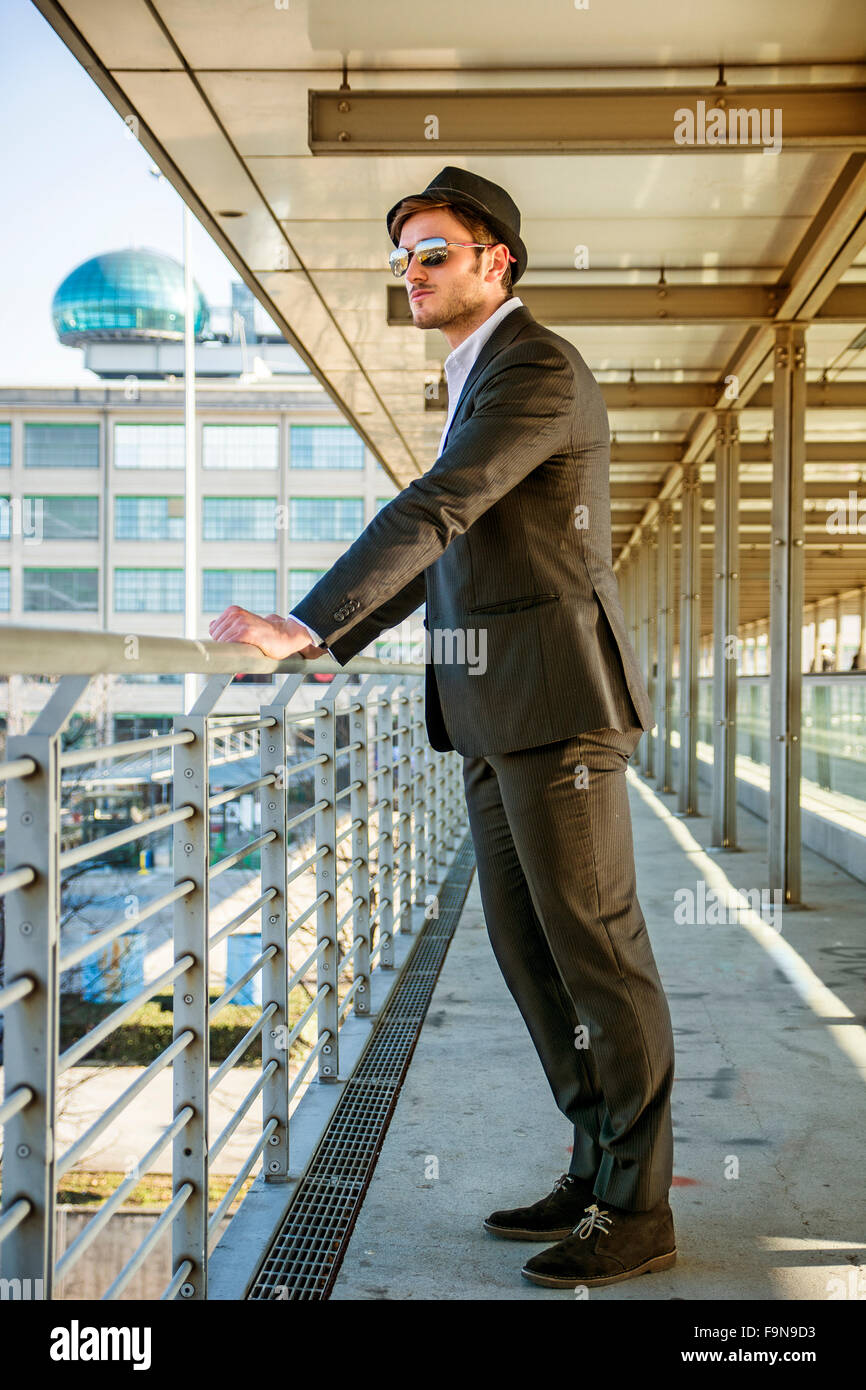 Voller Länge Portrait von Stylist junge Mann trägt Anzug und Hut auf der Seite von Balkon während stützte sich auf Handlauf in B Stockfoto