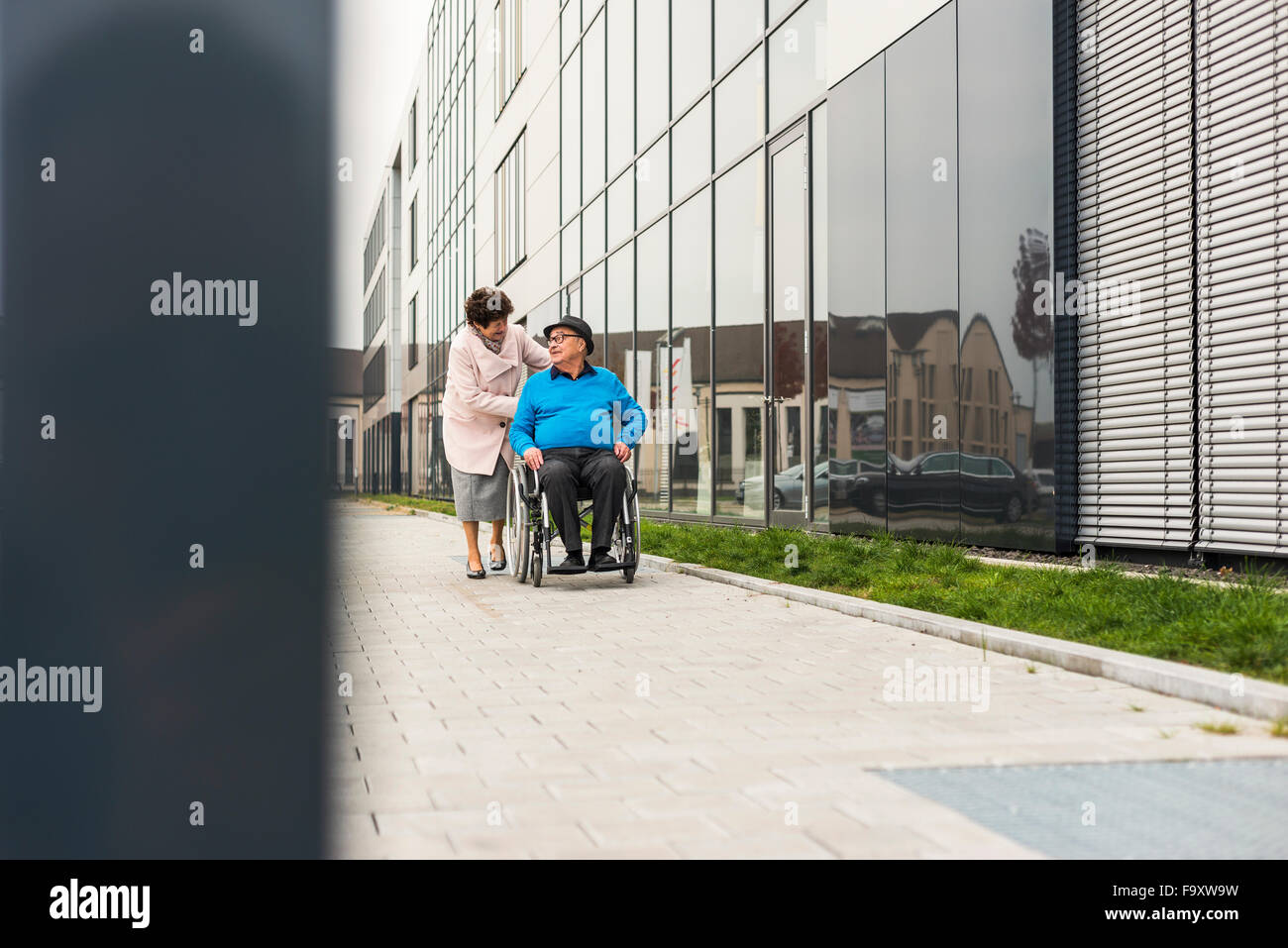 Ältere Frau, die Mann im Rollstuhl auf Bürgersteig schieben Stockfoto