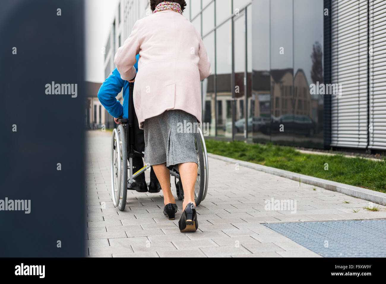 Ältere Frau, die Mann im Rollstuhl auf Bürgersteig schieben Stockfoto