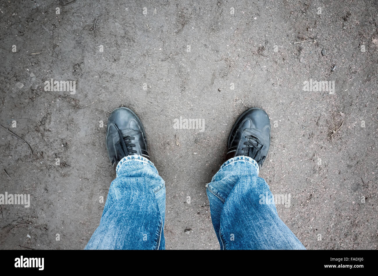 Männliche Füße in Blue Jeans und schwarze Schuhe stehen auf schmutzigen Landstraße, Egoperspektive Stockfoto