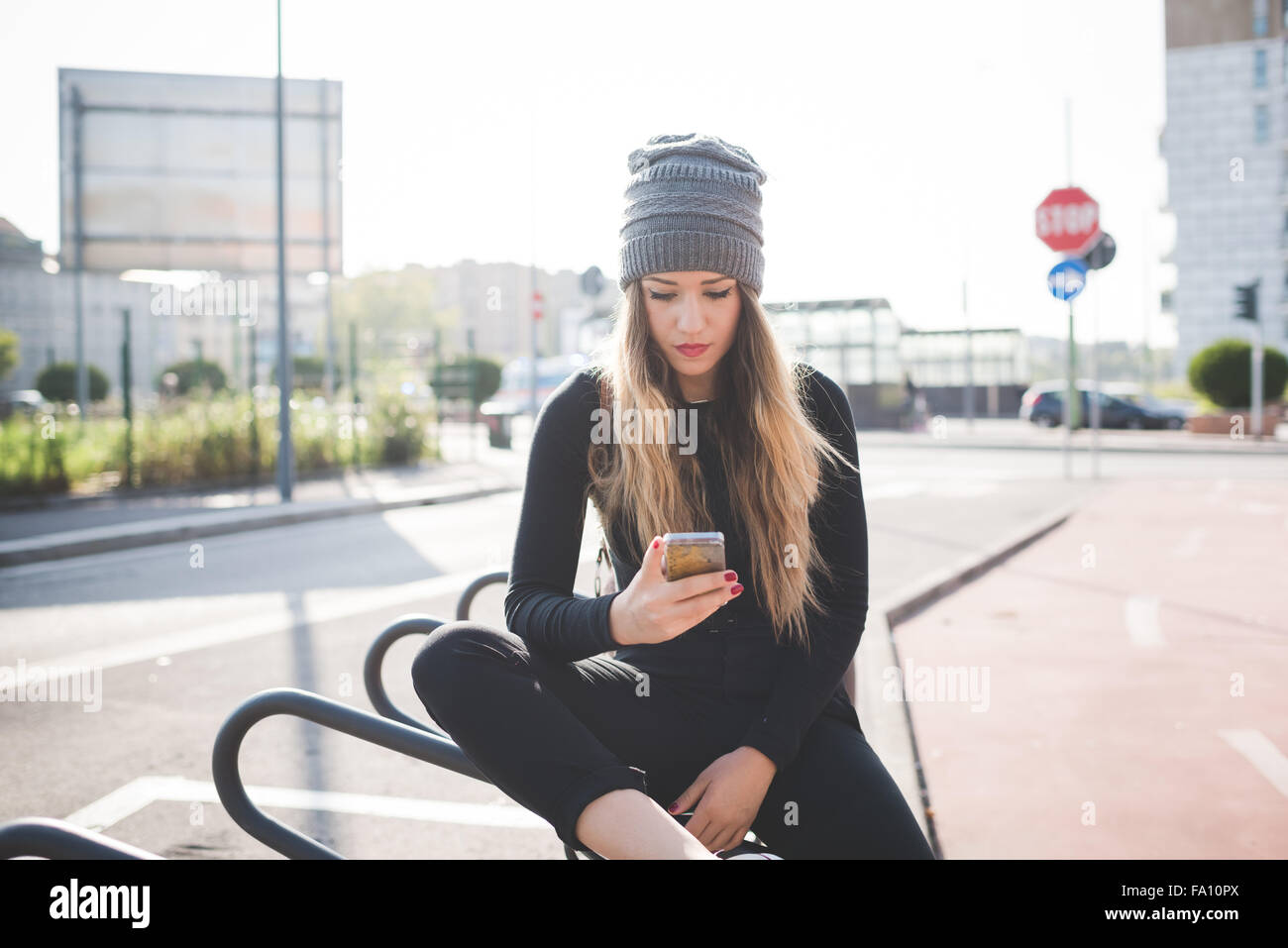 Knie Figur des jungen schönen kaukasischen blonde glatte Haare Frau sitzen im Freien in der Stadt, Smartphone Griffstück nach unten Tippen auf den Bildschirm - Kommunikation, Technologie, soziale Netzwerk-Konzept Stockfoto