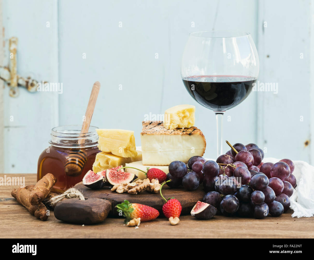 Glas Rotwein, Käse vom Brett, Trauben, Feigen, Erdbeeren, Honig und Brot klebt am rustikalen Holztisch, blauer Hintergrund, sele Stockfoto