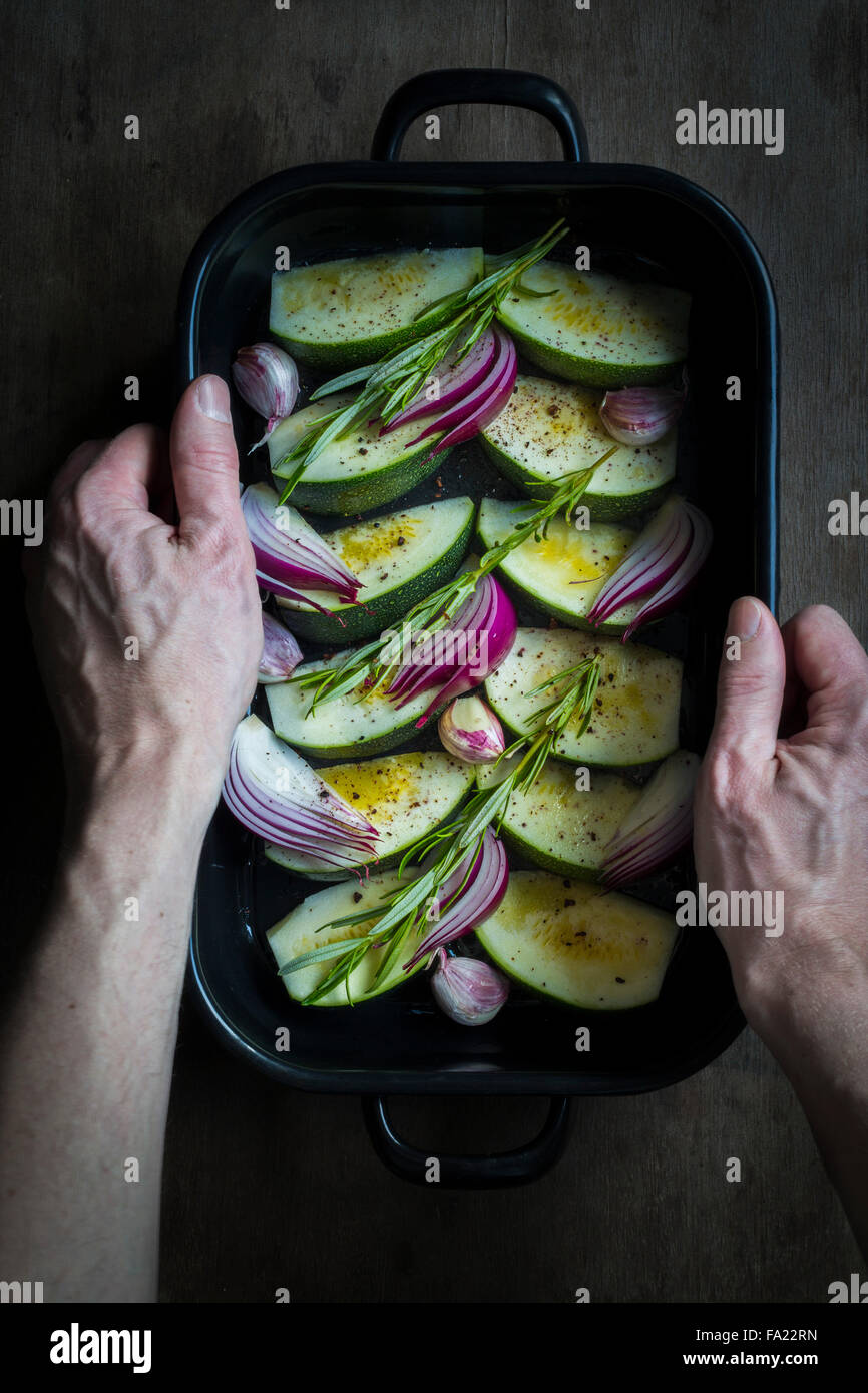 Hände halten eine Auflaufform mit Zucchini, Zwiebeln und frischen Kräutern zum Kochen vorbereitet Stockfoto