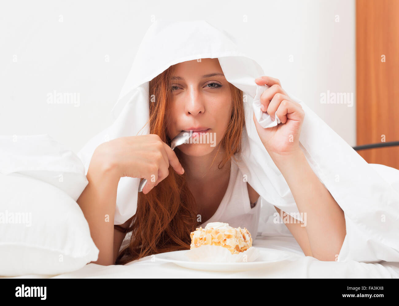 Junge Frau, die süßen Kuchen unter Laken im Bett Essen Stockfoto