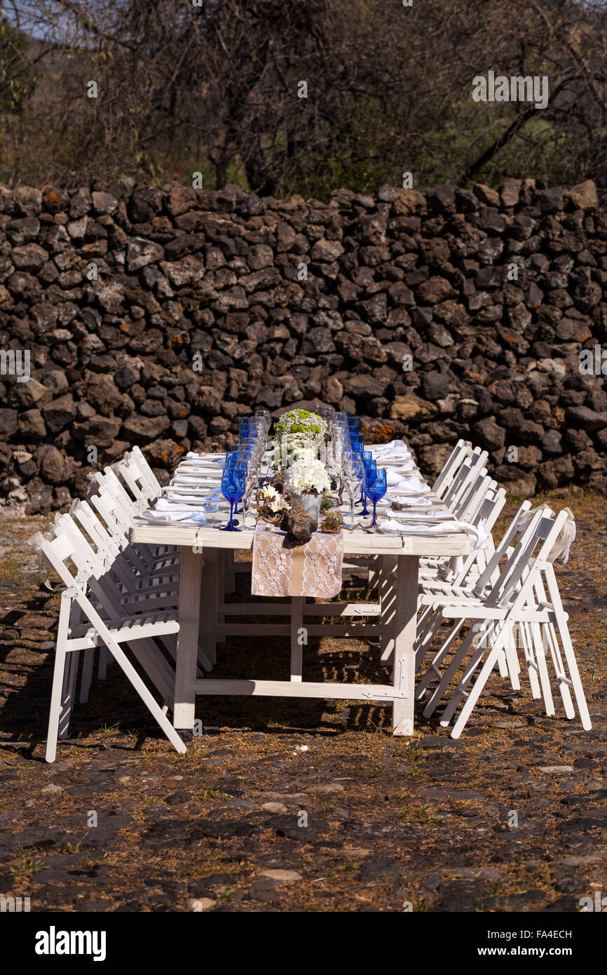 Weißen Tisch und hölzerne Stühle set mit blauen Gläsern, Blumen und Dekorationen für eine Feier im Freien. Stockfoto