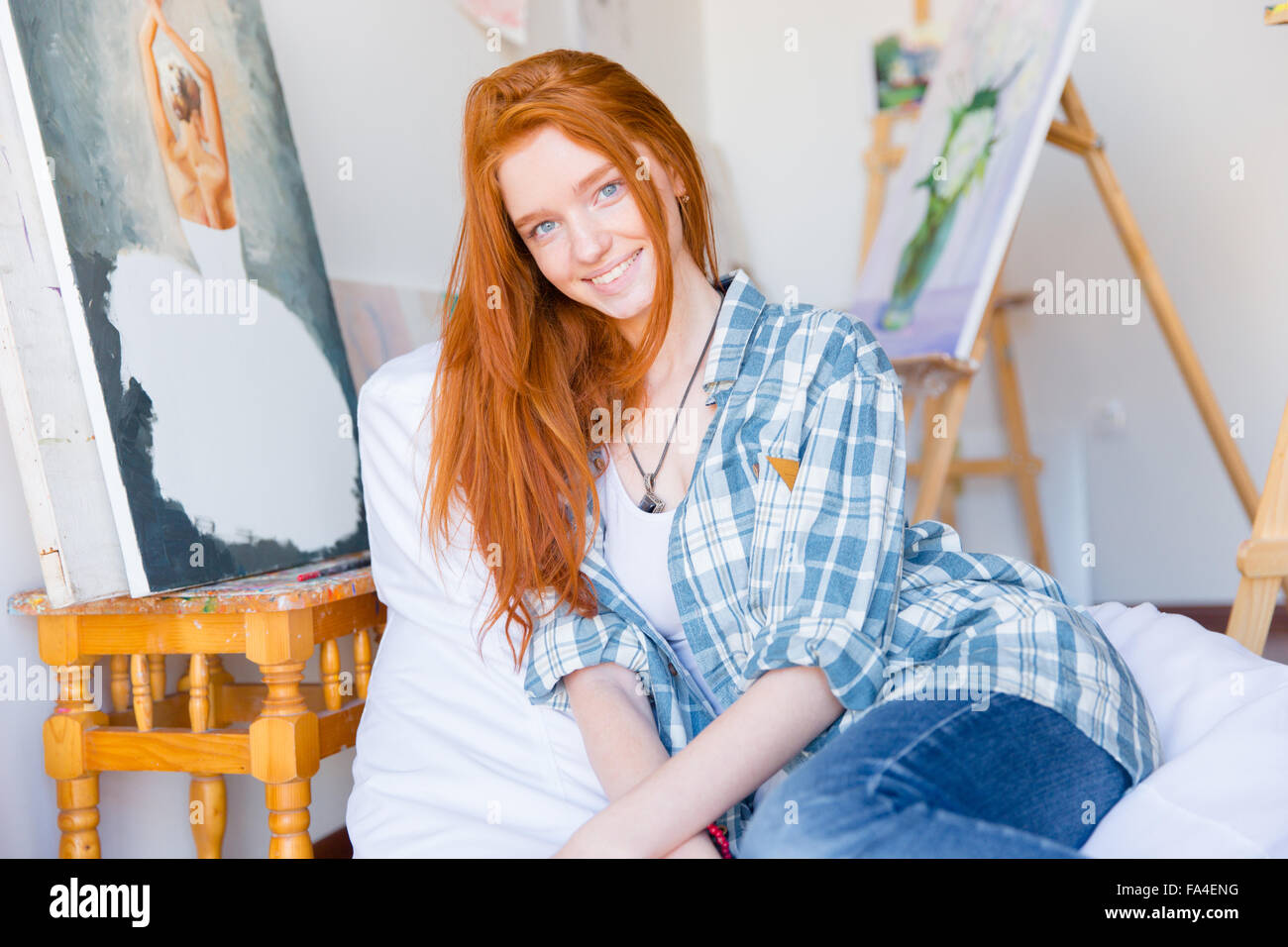 Attraktive glückliche junge Frau mit langen roten Haaren im karierten Hemd und Jeans sitzen auf weichen weißen Sitzsack in Kunstwerkstatt Stockfoto