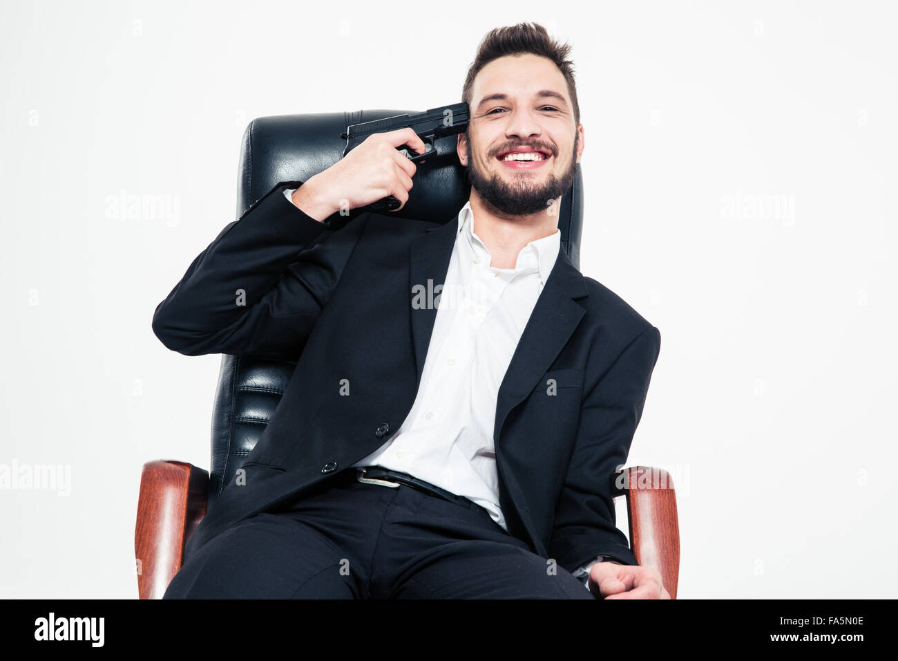 Lächeln auf den Lippen schön bärtigen Business-Mann im schwarzen Anzug und weißem Hemd sitzen im Bürostuhl mit Pistole an der Schläfe über weiß ba Stockfoto