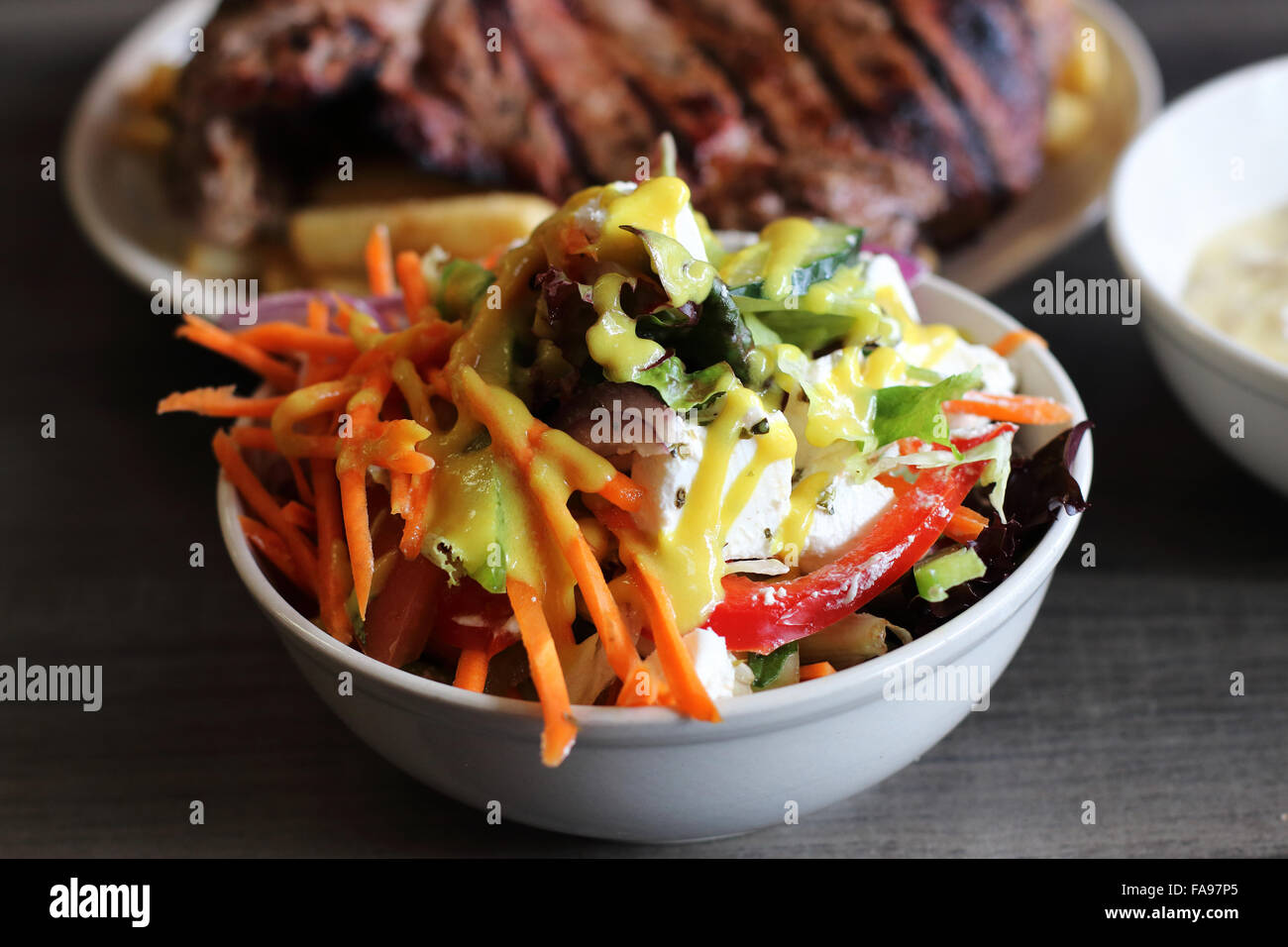 Eine Schüssel mit griechischer Salat in eine weiße Schüssel mit Steak und Chips im Hintergrund Stockfoto