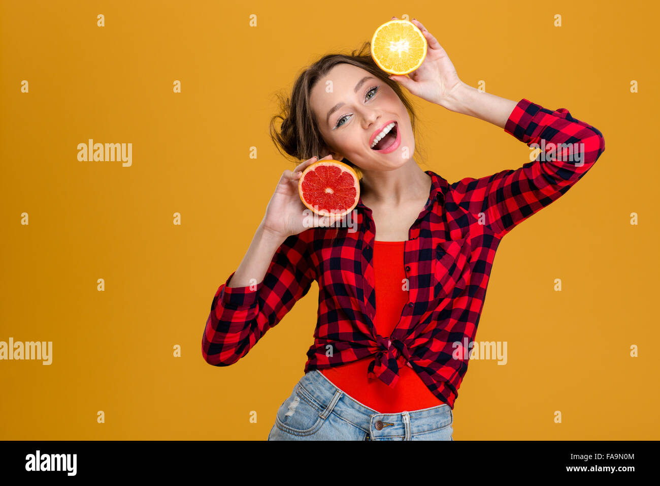 Freudige schöne junge Frau im karierten Hemd hält Hälften von Zitrusfrüchten über gelbem Hintergrund Stockfoto