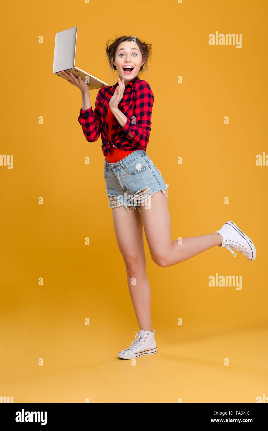 Gesamte Länge der süße schöne fröhliche junge Frau im karierten Hemd und Jeans Shorts posiert mit Laptop auf gelbem Hintergrund Stockfoto