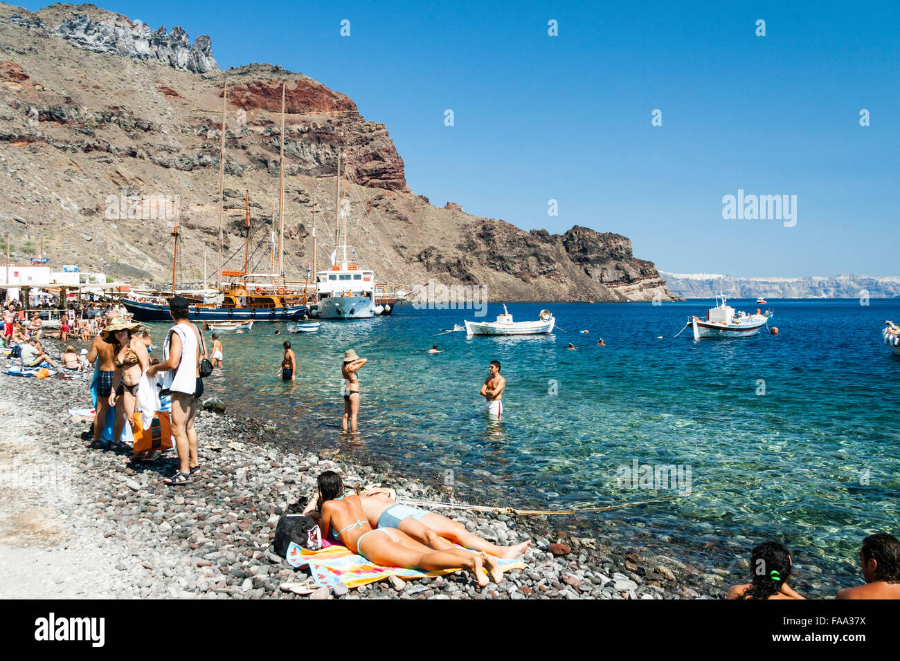 Santorini, Korfos, Aussicht auf schmalen Kieselstrand, mit Touristen und Sonnenanbeter paddeln und am Strand. Blaues Meer, kleine Boote und felsigen Klippen. Stockfoto