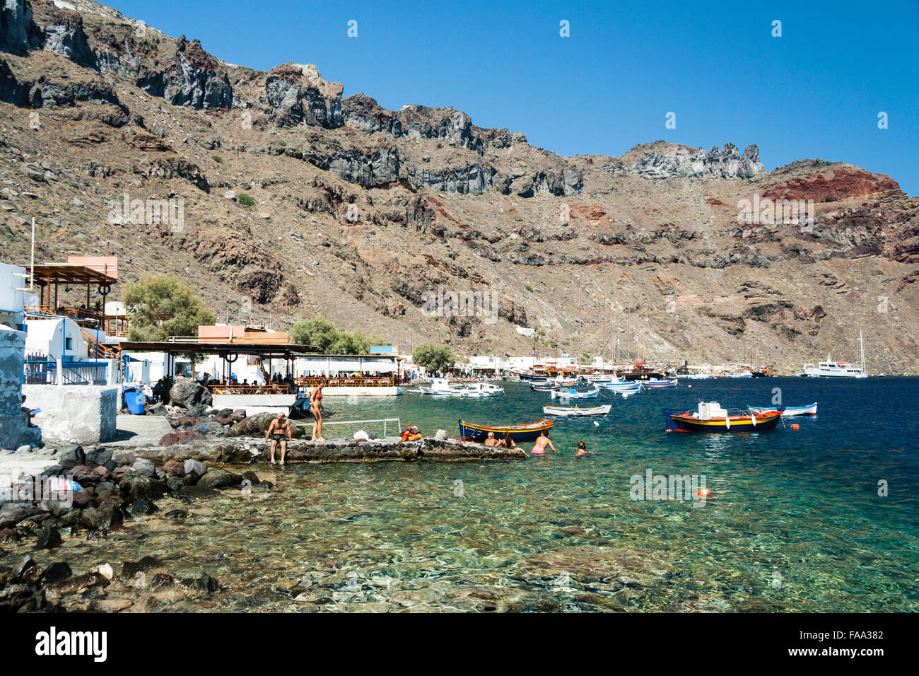 Santorini, Korfos, Aussicht auf schmalen Kieselstrand, mit Touristen und Sonnenanbeter paddeln und am Strand. Blaues Meer, kleine Boote und felsigen Klippen. Stockfoto