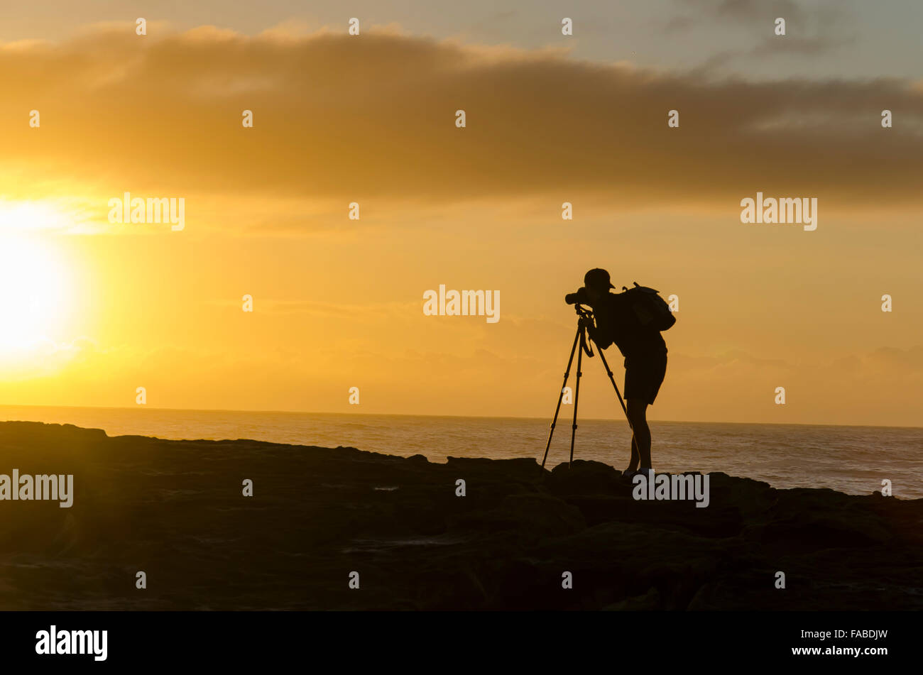 Ein junger männlicher Fotograf wird in der Dämmerung auf den Felsen in der Nähe eines australischen Strands in Sydney gegen die aufgehende Sonne geschildet Stockfoto