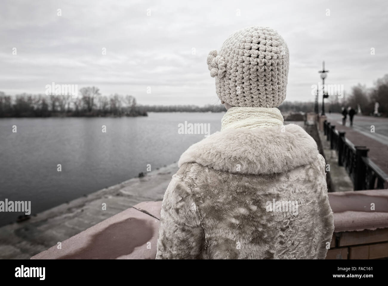 Eine Frau mit einer Mütze und einem Pelzmantel blickt auf den Fluss in einem kalten und traurigen grauer Wintermorgen Stockfoto