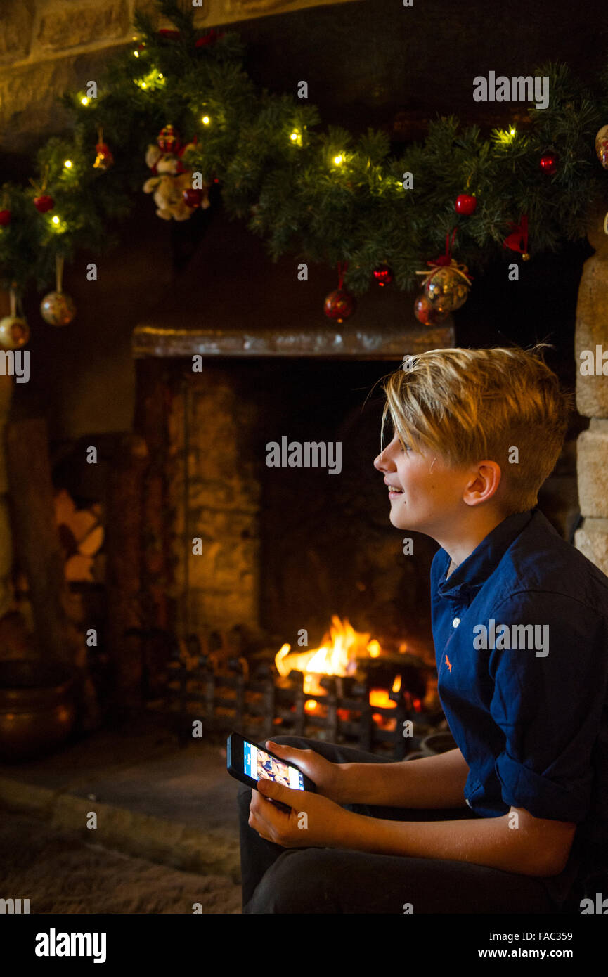 Ein kleiner Junge zu Weihnachten mit seinem neuen Smartphone vor dem offenen Feuer und Weihnachtsschmuck Stockfoto