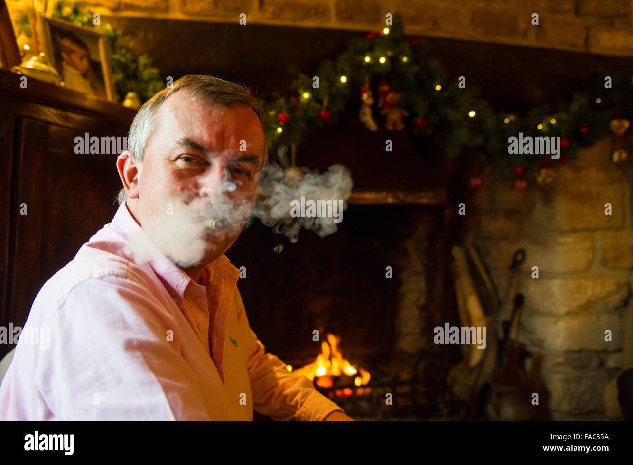 Ein Mann raucht eine Zigarre vor dem offenen Feuer in einem Landhaus zu Weihnachten Stockfoto