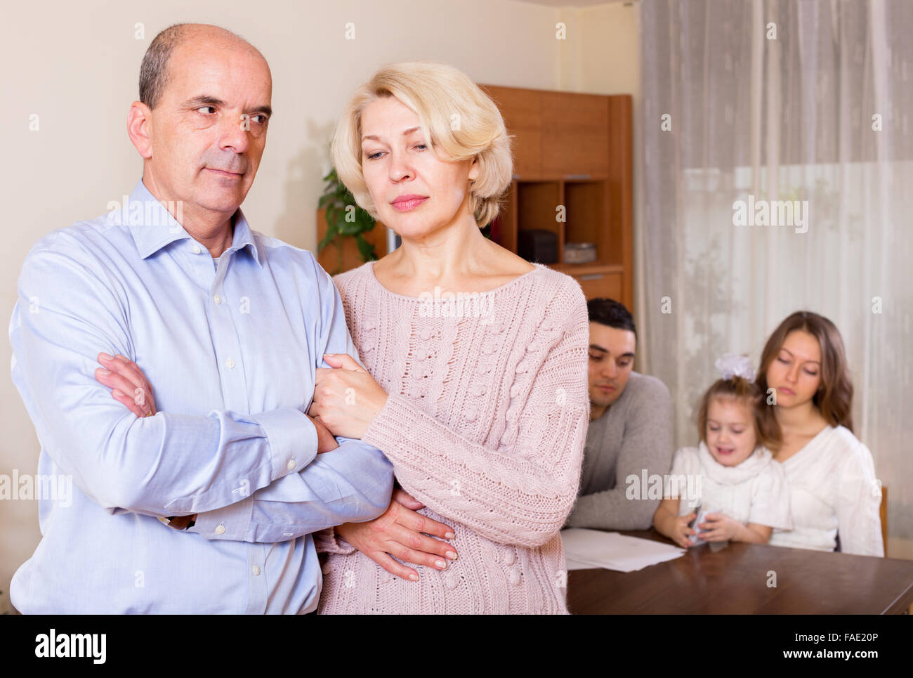 Traurig älteres Paar auf Hintergrund junger Eltern mit Kind zu Hause Stockfoto