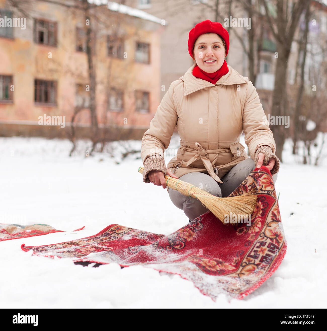 Lächelnde Frau in roten Mütze reinigt Teppich mit Schnee im Wintertag im freien Stockfoto