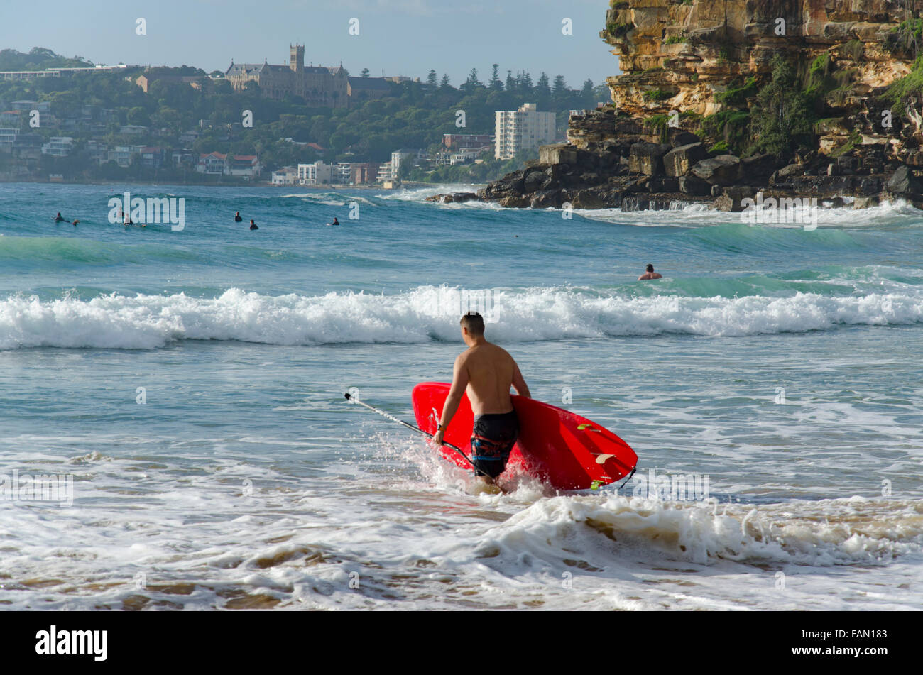 Ein Mann mit einem roten Stand-up-Paddleboard (SUP) steigt an einem sonnigen Sydney-Morgen in Australien in die Brandung am Freshwater Beach ein Stockfoto