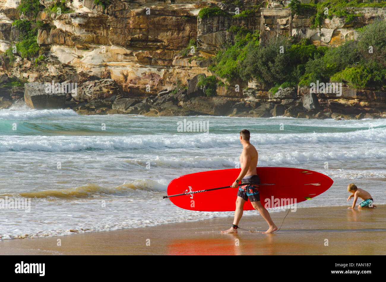 Ein Mann mit einem roten Stand-up-Paddleboard (SUP) steigt an einem sonnigen Morgen in Sydney in die Brandung am Freshwater Beach ein Stockfoto