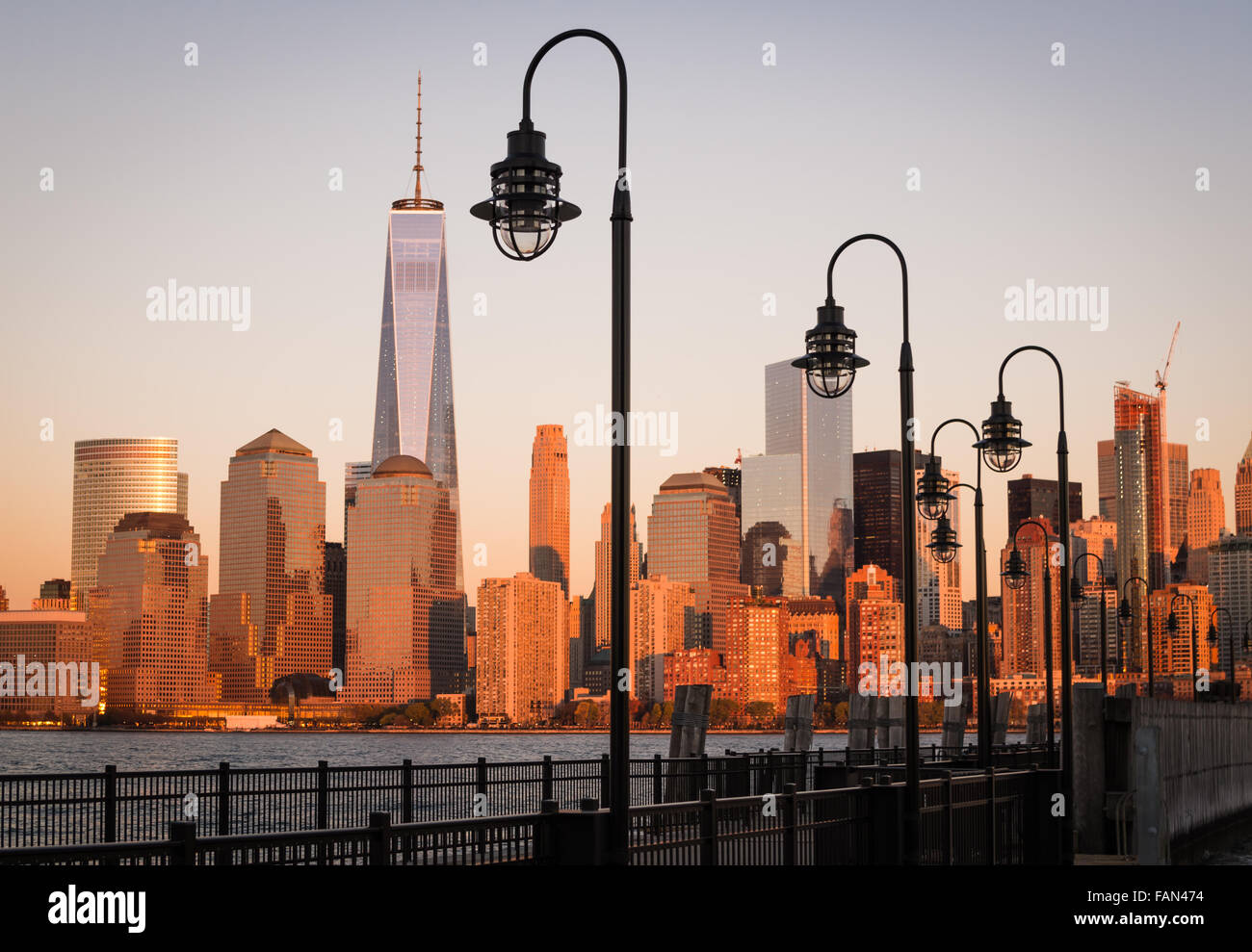 Ein verlassener Pier mit Straßenlaternen freut über den Hudson River auf die unteren Skyline von Manhattan, die den goldenen Sonnenuntergang widerspiegelt. Stockfoto