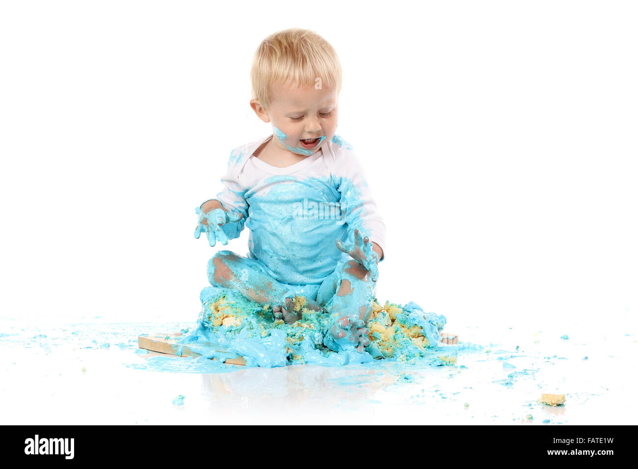Ein einjähriges Baby junge Zerschlagung einer blauen Geeiste Geburtstagstorte auf einem Holzbrett. Bild wird auf einem weißen Hintergrund isoliert. Stockfoto