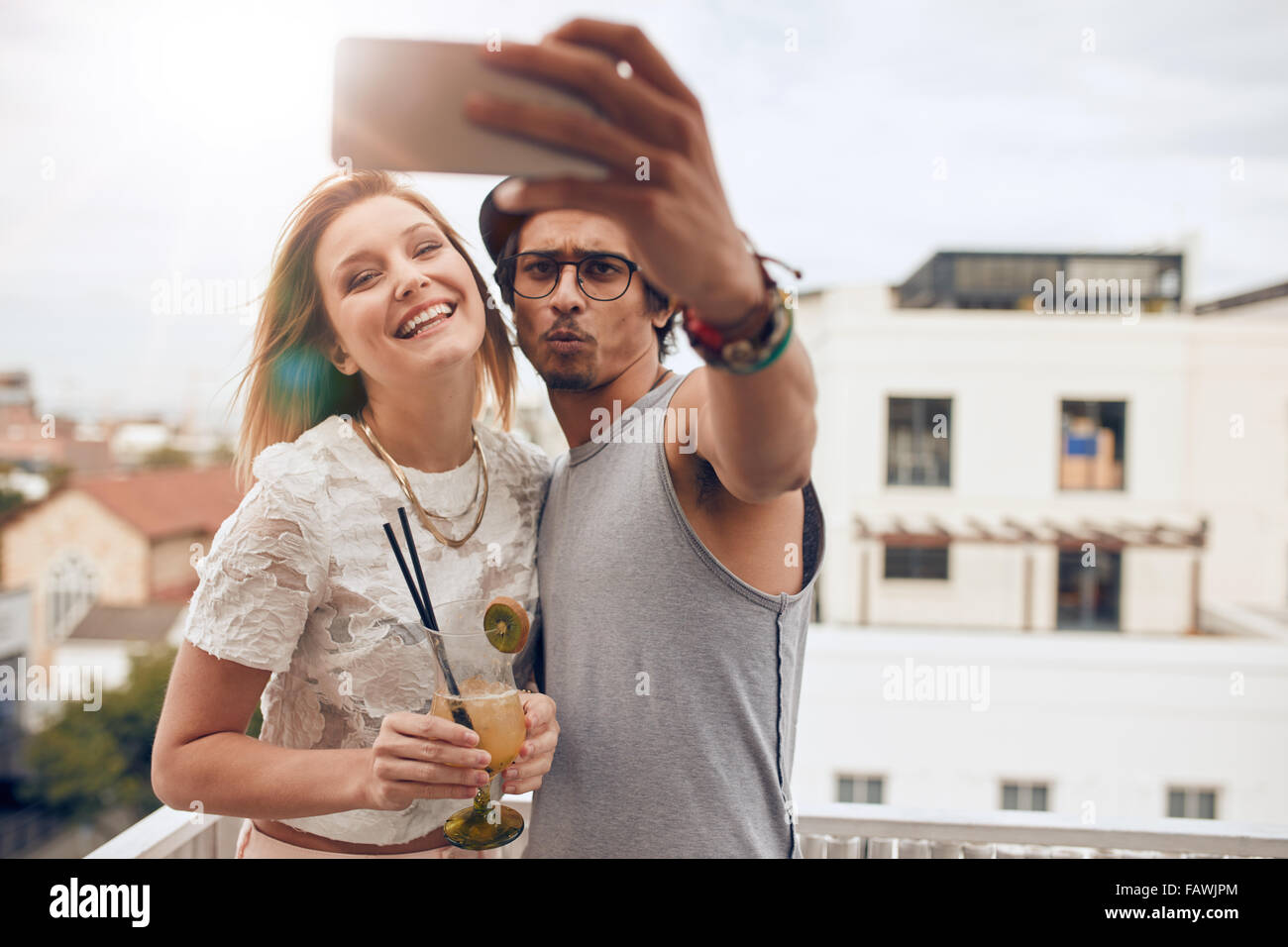 Zwei junge Freunde, die auf dem Dach eine Selfie übernehmen. Mann hält Smartphone und Selbstportrait mit Frau hält einen cocktail dur Stockfoto