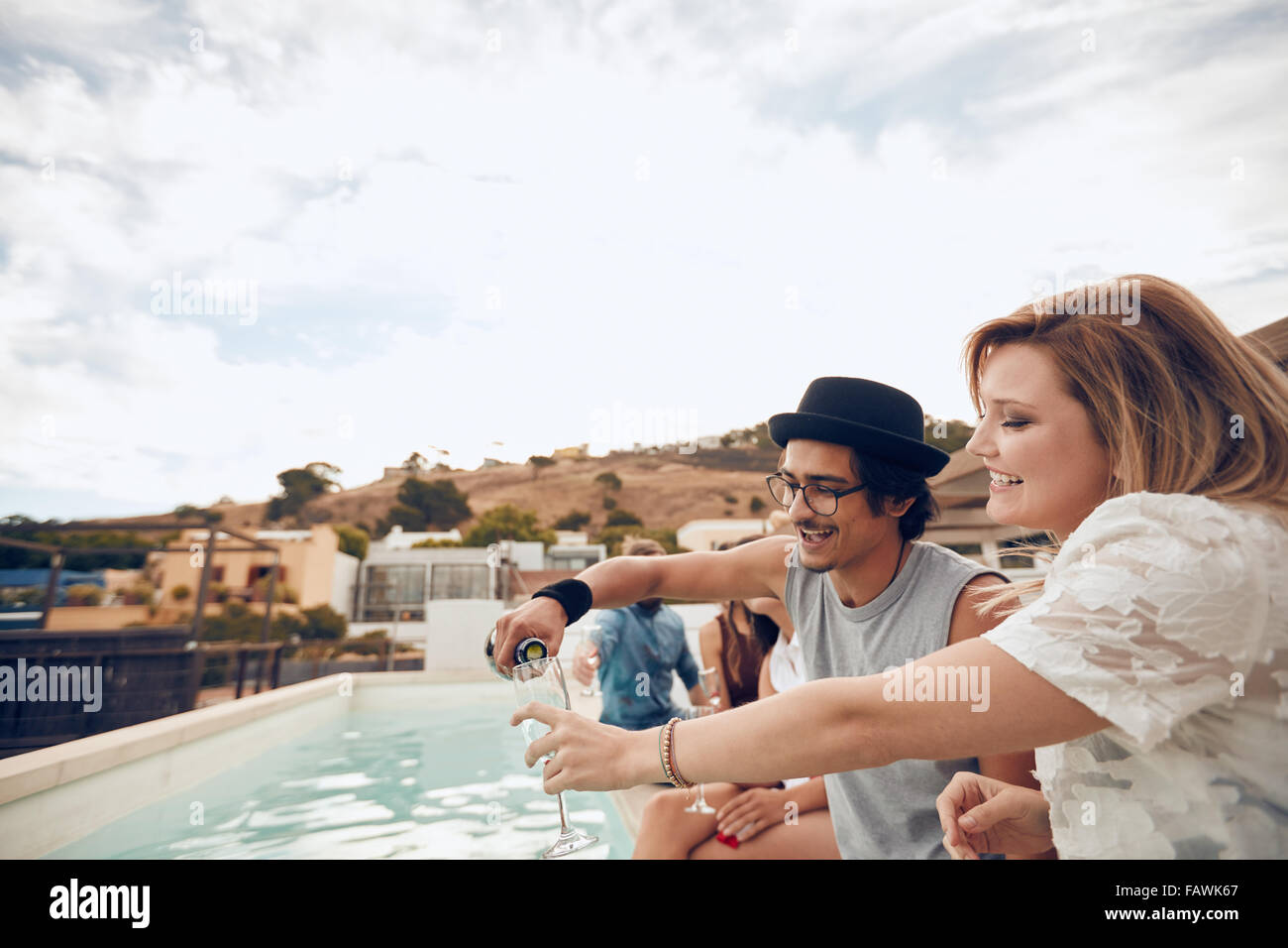Junger Mann gießt Champagner im Glas von seiner Freundin während der Party am Pool. Junge Menschen, die Spaß am Dach p Stockfoto