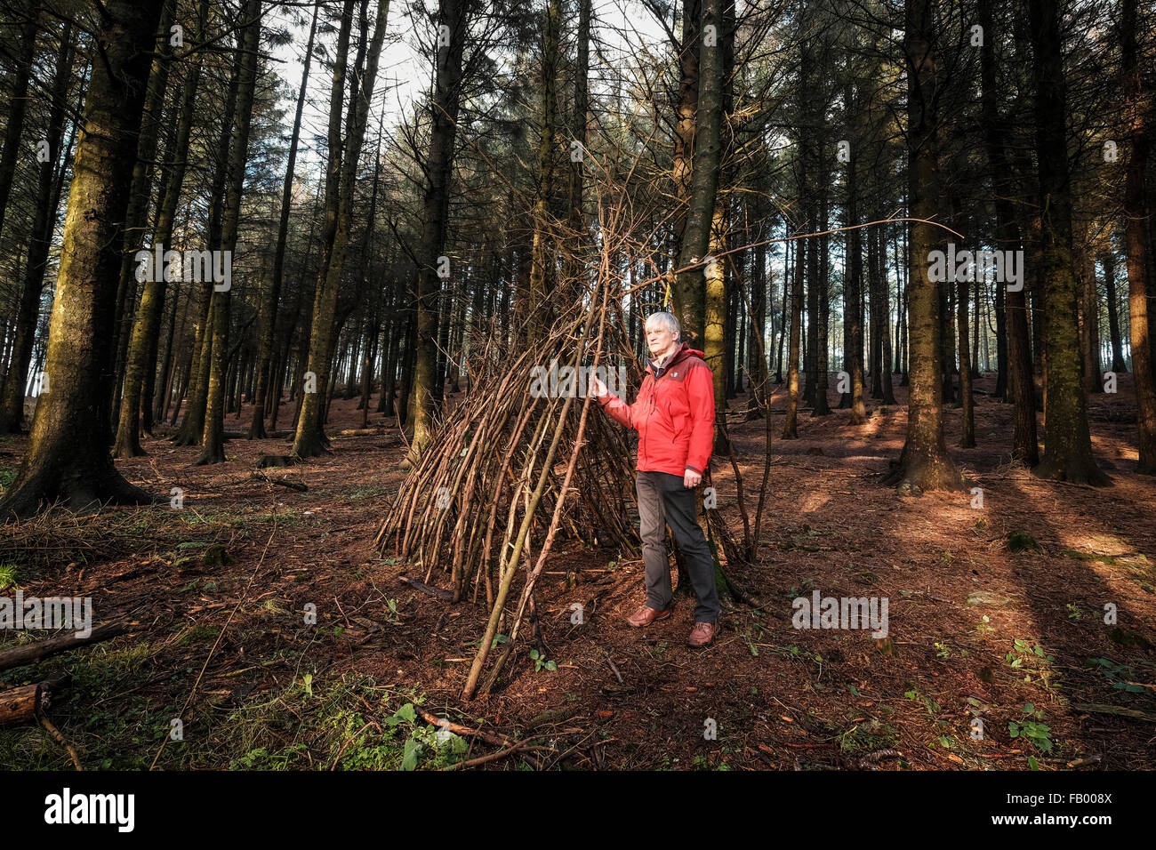 Leuchtfeuer fiel, Lancashire, England. Mann in rot Outdoorjacke neben dem teilweise Stand gebaut Tierheim gemacht aus Stöcken und Zweigen. Stockfoto