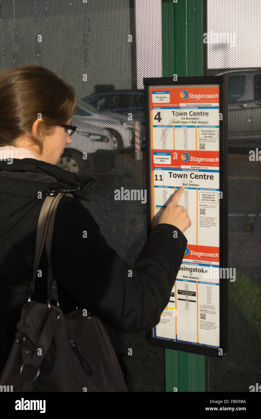 Eine junge Frau in ihren Zwanzigern mit Mantel und Handtasche, die einen Busfahrplan konsultiert und auf 'Town Center' an einer Bushaltestelle, Basingstoke, Großbritannien, zeigt Stockfoto