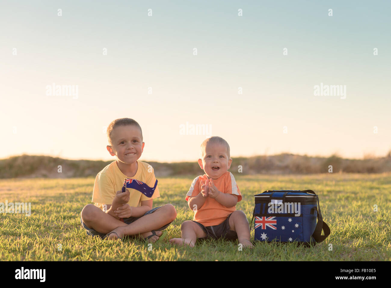 Lächelnde Kinder sitzen auf dem Rasen, bei Sonnenuntergang und mit australischer Flagge und Kühlbox Stockfoto