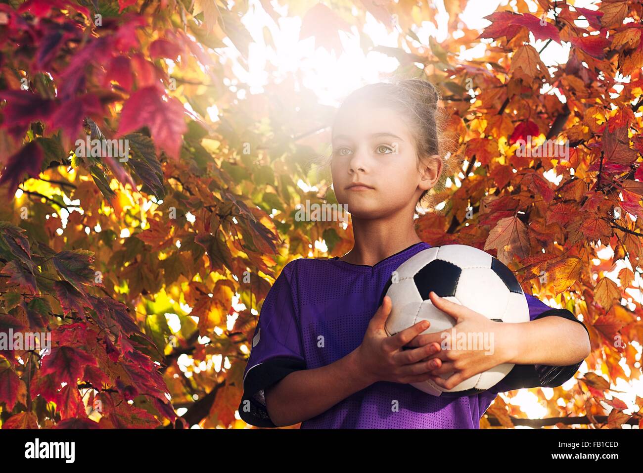 Mädchen mit Baum bedeckt im Herbst Blätter halten Fußball wegschauen Stockfoto