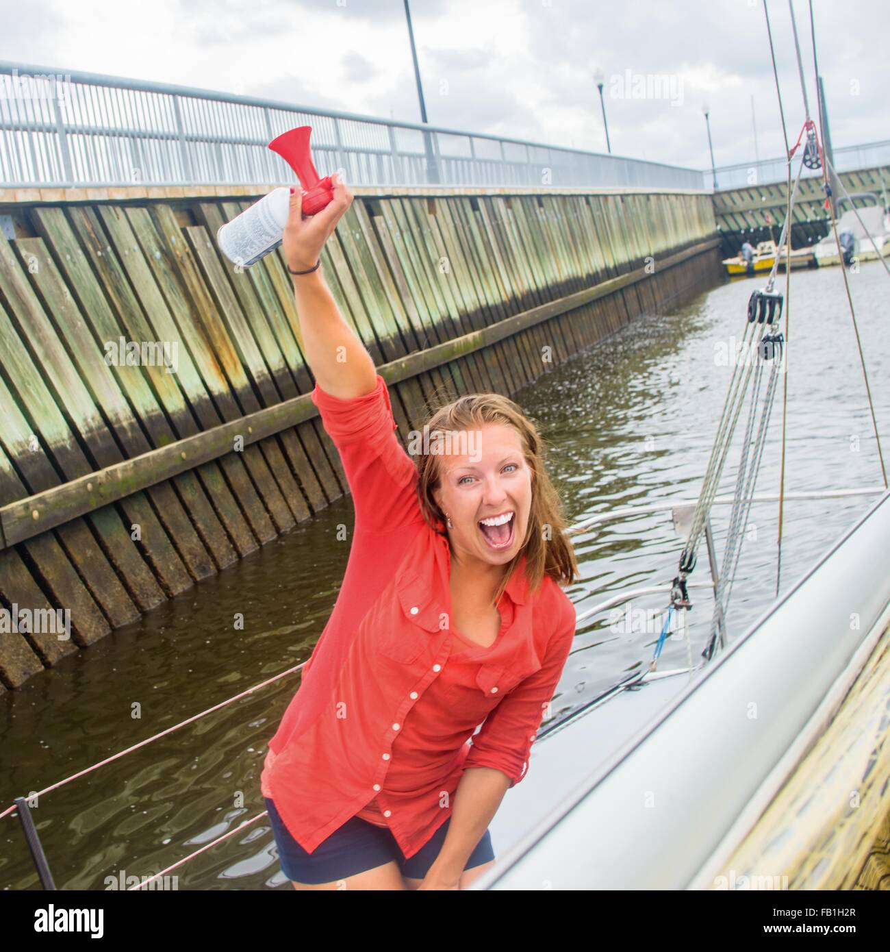 Junge Frau auf Segelboot in Marina Arm angehoben halten Lufthorn, Blick in die Kamera geöffnet Mund Lächeln Stockfoto