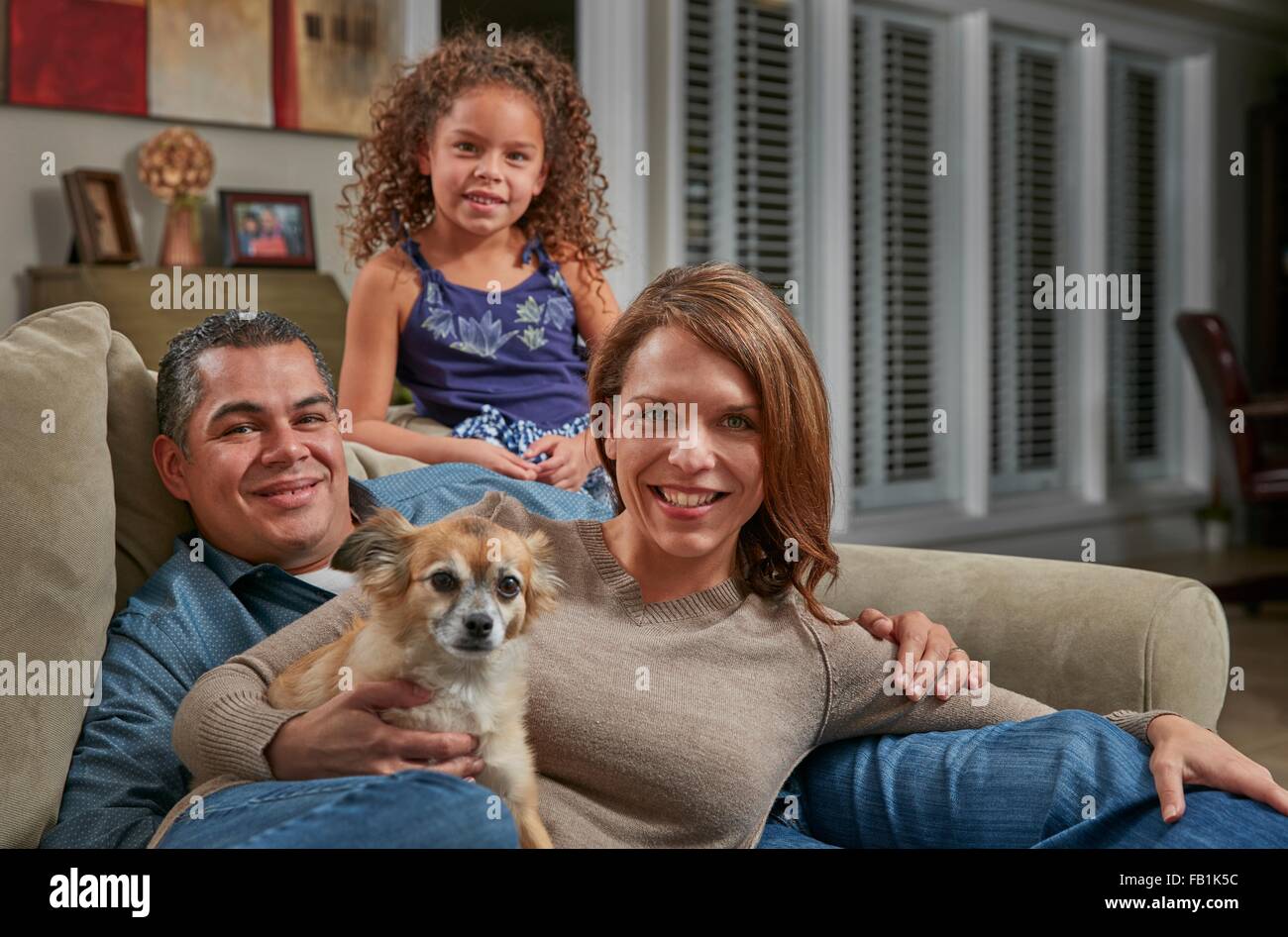 Eltern und Mädchen im Wohnzimmer auf dem Sofa mit Blick auf die Kamera zu Lächeln Hund Stockfoto