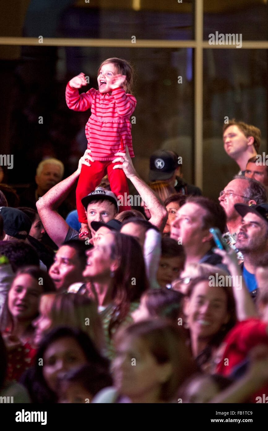 Oberhalb der gemischtrassigen Masse auf Vaters Schultern, Uhren ein aufgeregt Kleinkind eine Bühne auf einem Christmas Festival in einem Einkaufszentrum in San Clemente, CA, zu zeigen. Stockfoto